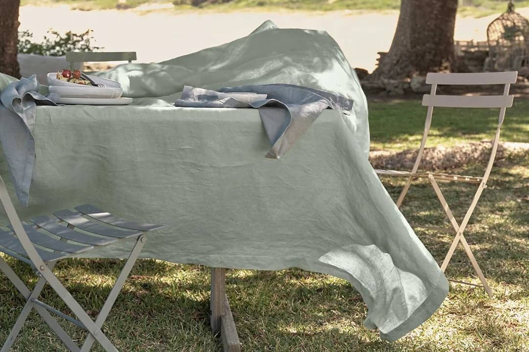 Sage green linen tablecloth draped over an outdoor dining table, showing natural texture and relaxed drape in sunlight