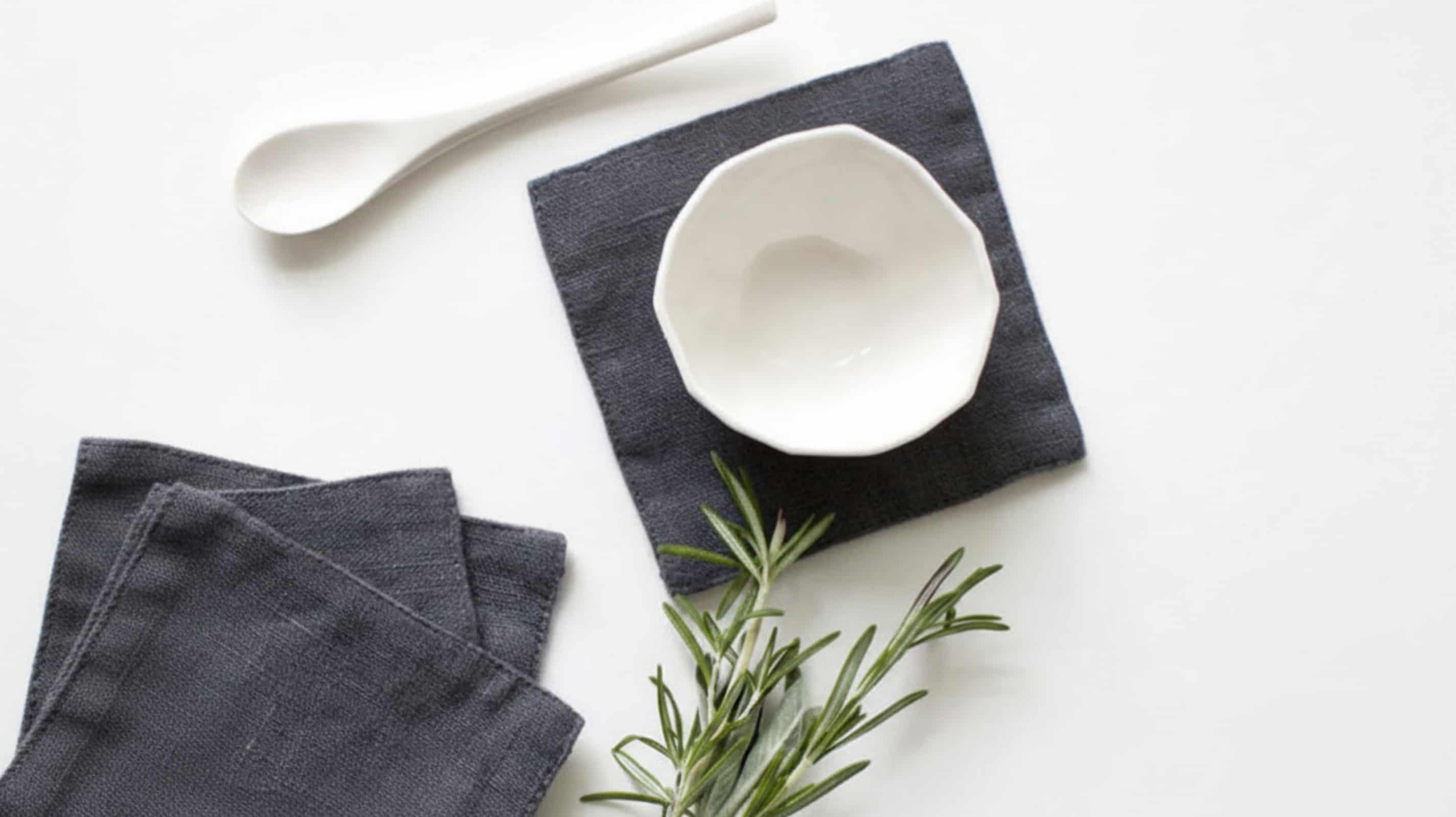 Dark grey washed linen coasters with white ceramic bowl, spoon and fresh rosemary on white background, flat lay