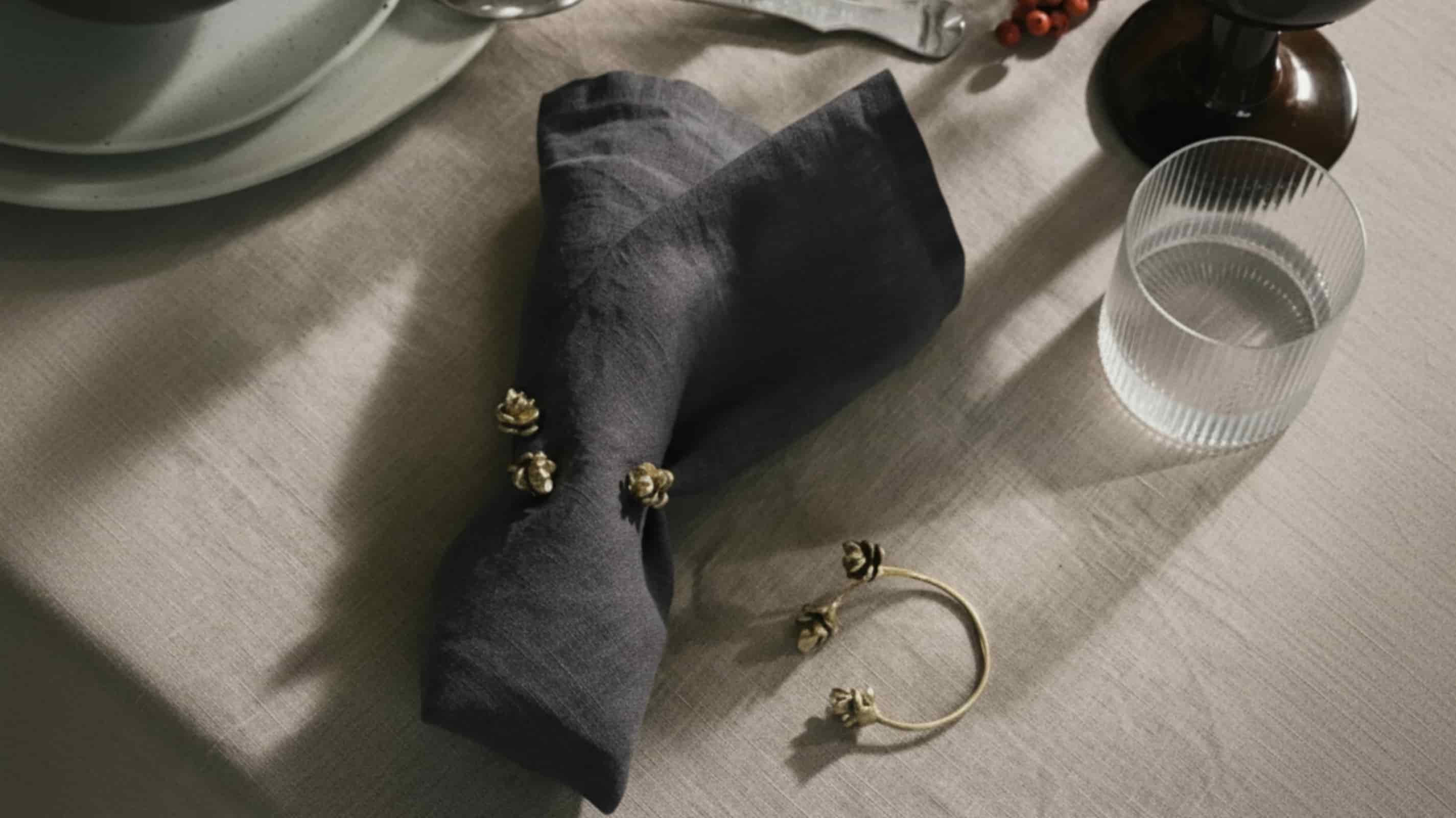Dark grey washed linen napkin folded with gold floral napkin ring on natural linen placemat with ribbed water glass and dark berry vase