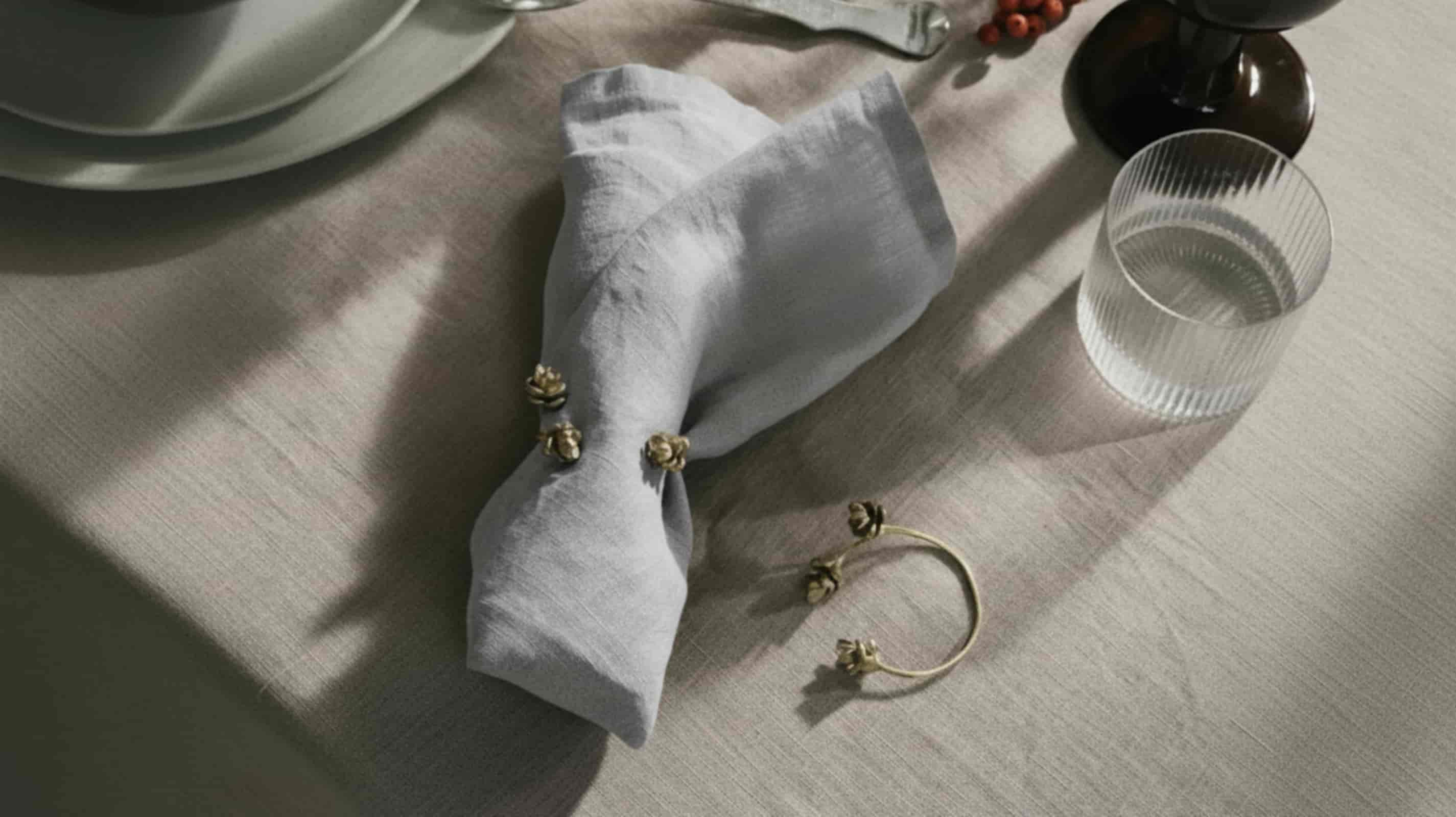Light grey washed linen napkin folded with gold floral napkin ring on natural linen placemat with ribbed water glass and dark berry vase