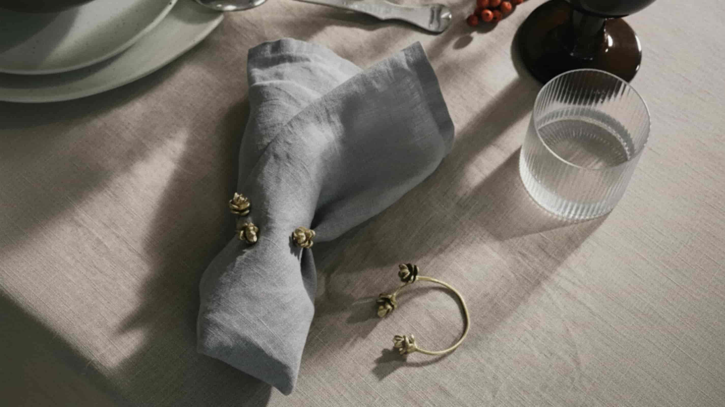 Middle grey washed linen napkin folded with gold floral napkin ring on natural linen placemat with ribbed water glass and dark berry vase