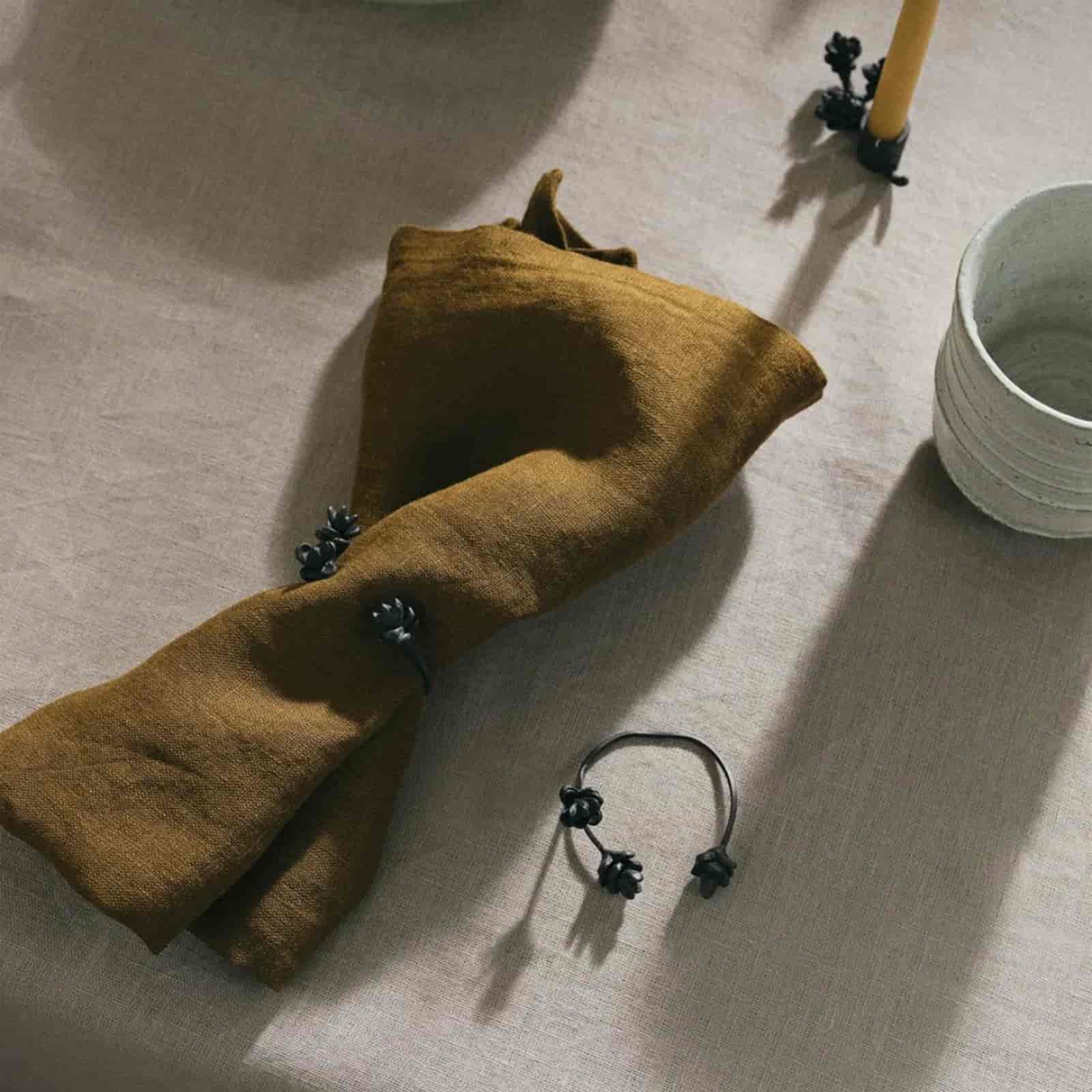 Mustard yellow washed linen napkin loosely tied with black floral napkin ring on natural linen tablecloth with beeswax candle and white ceramic bowl