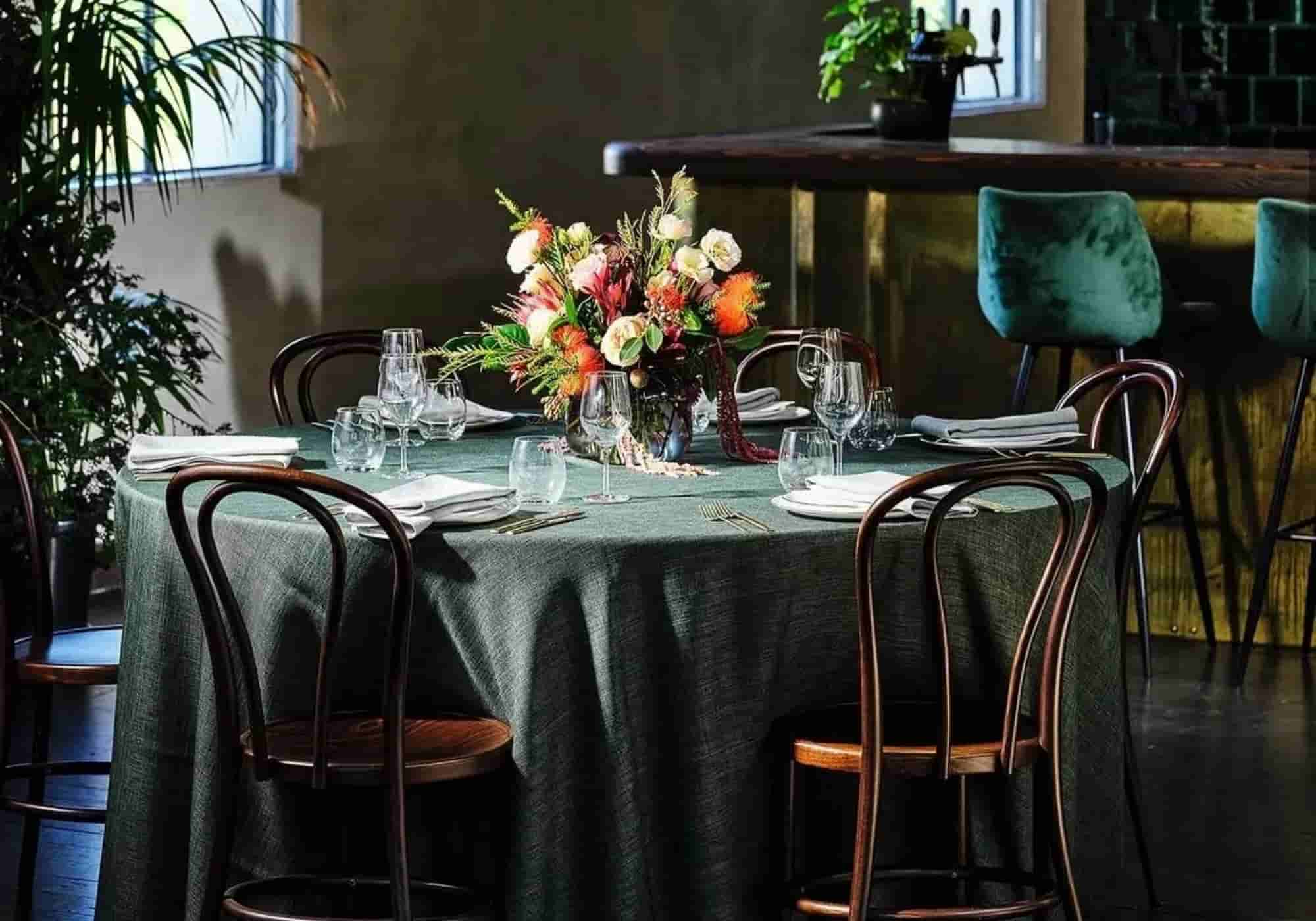 Sage green linen round tablecloth on a restaurant table with wild floral centerpiece, white linen napkins and bentwood chairs beside a bar counter