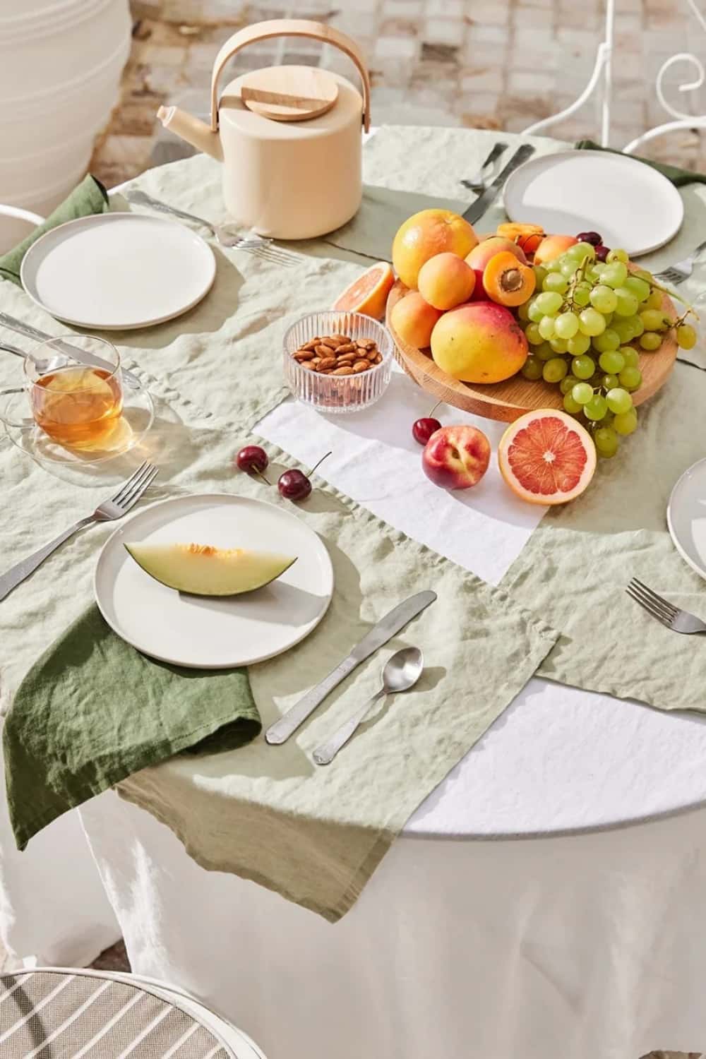 Sage green washed linen placemats on outdoor round table styled with fresh fruit, white plates and tea pot