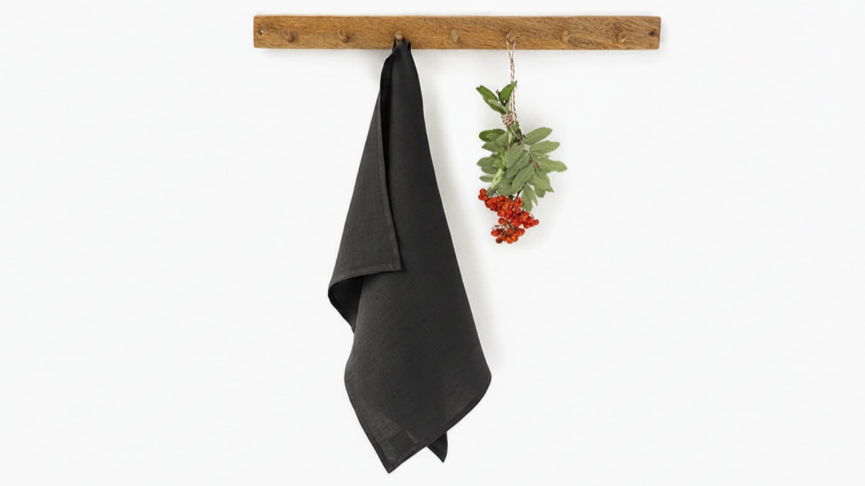 Dark grey washed linen tea towel hanging on rustic wooden hook rack with red berries, white background