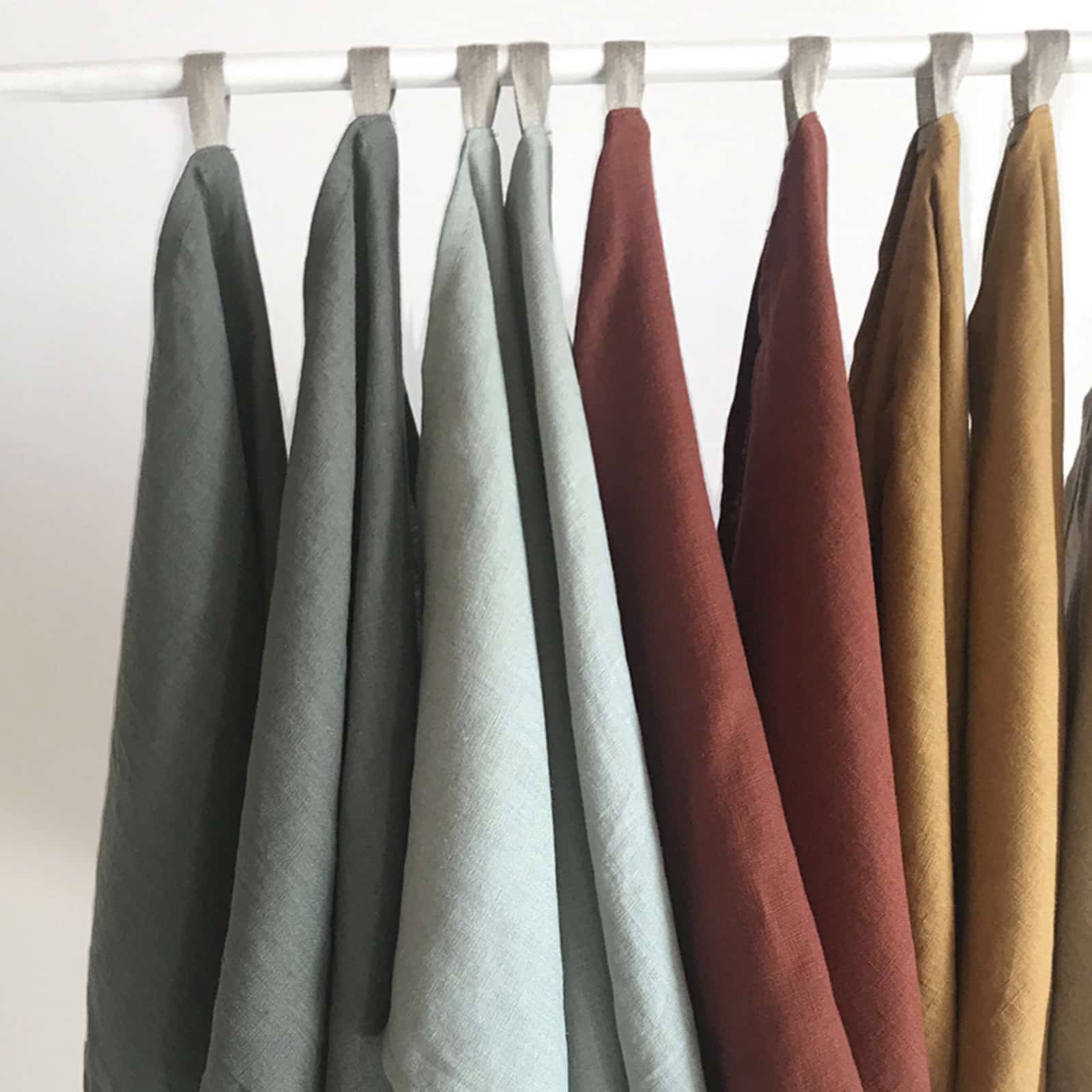 Multiple washed linen tea towels in sage green, light blue, burgundy, rust and mustard hanging on rod, color options display