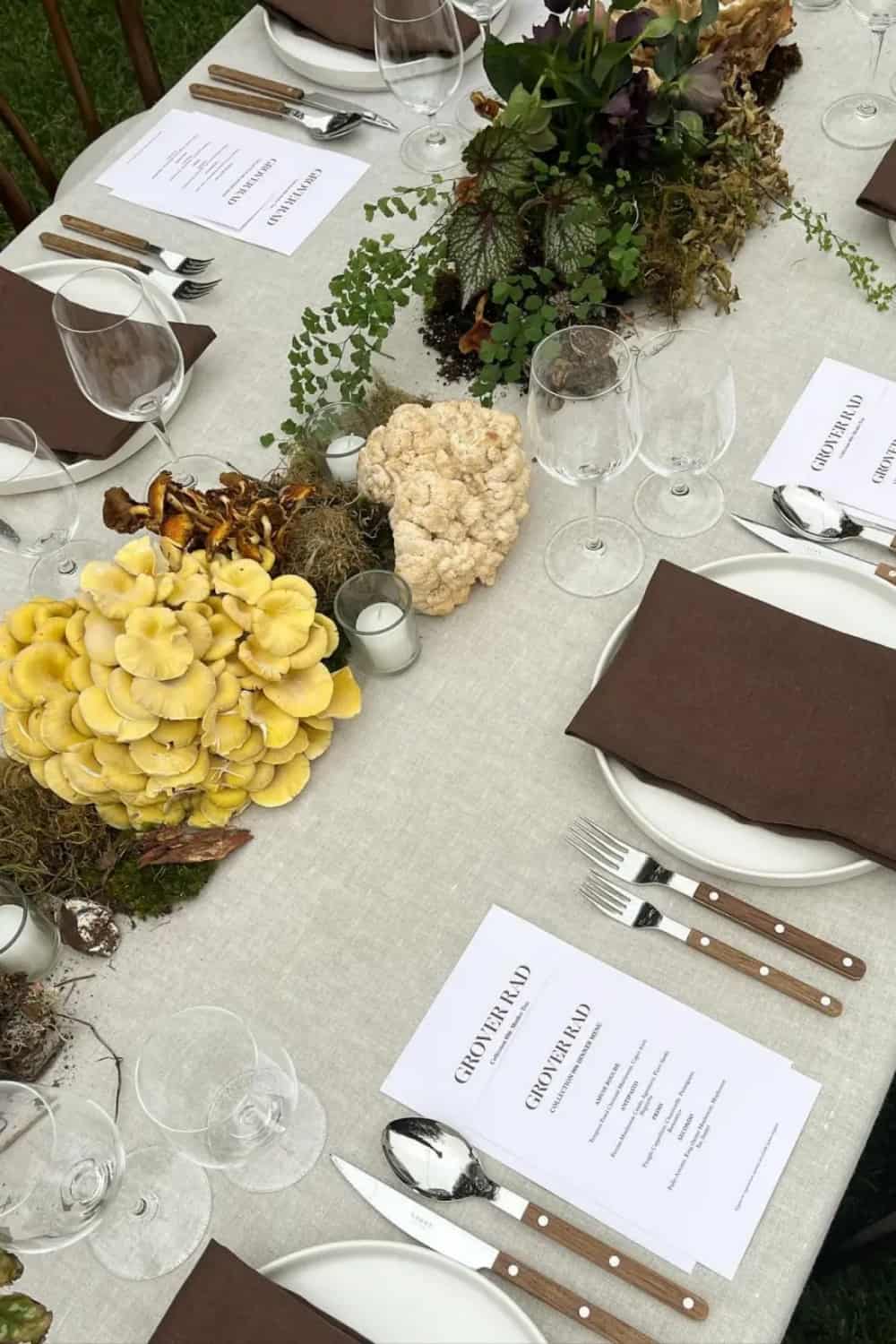 Overhead view of a white linen tablecloth outdoor dinner table with wild mushroom and botanical centerpiece, brown linen napkins and printed menu cards