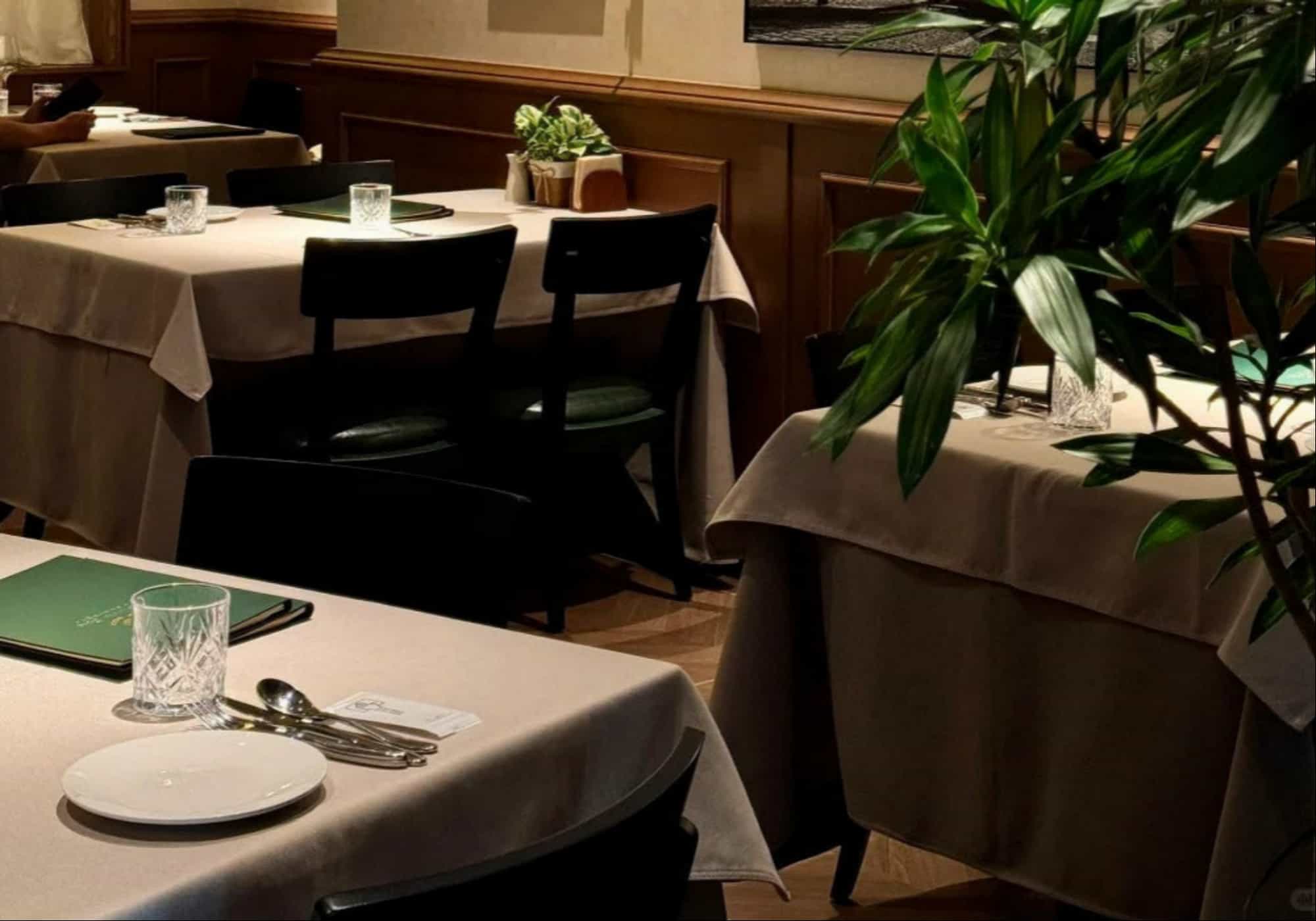 Modern restaurant interior with beige linen tablecloth, black chairs, crystal glasses and green plants in warm ambient lighting