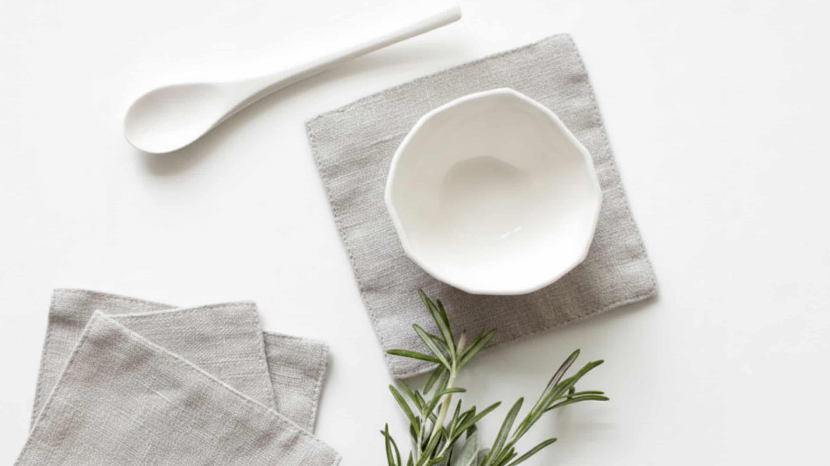 Light grey washed linen coasters with white ceramic bowl, spoon and fresh rosemary on white background, flat lay