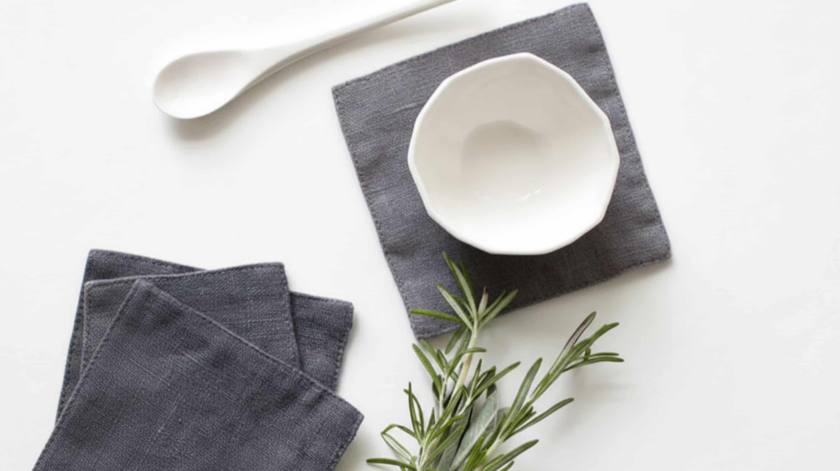 Middle grey washed linen coasters with white ceramic bowl, spoon and fresh rosemary on white background, flat lay