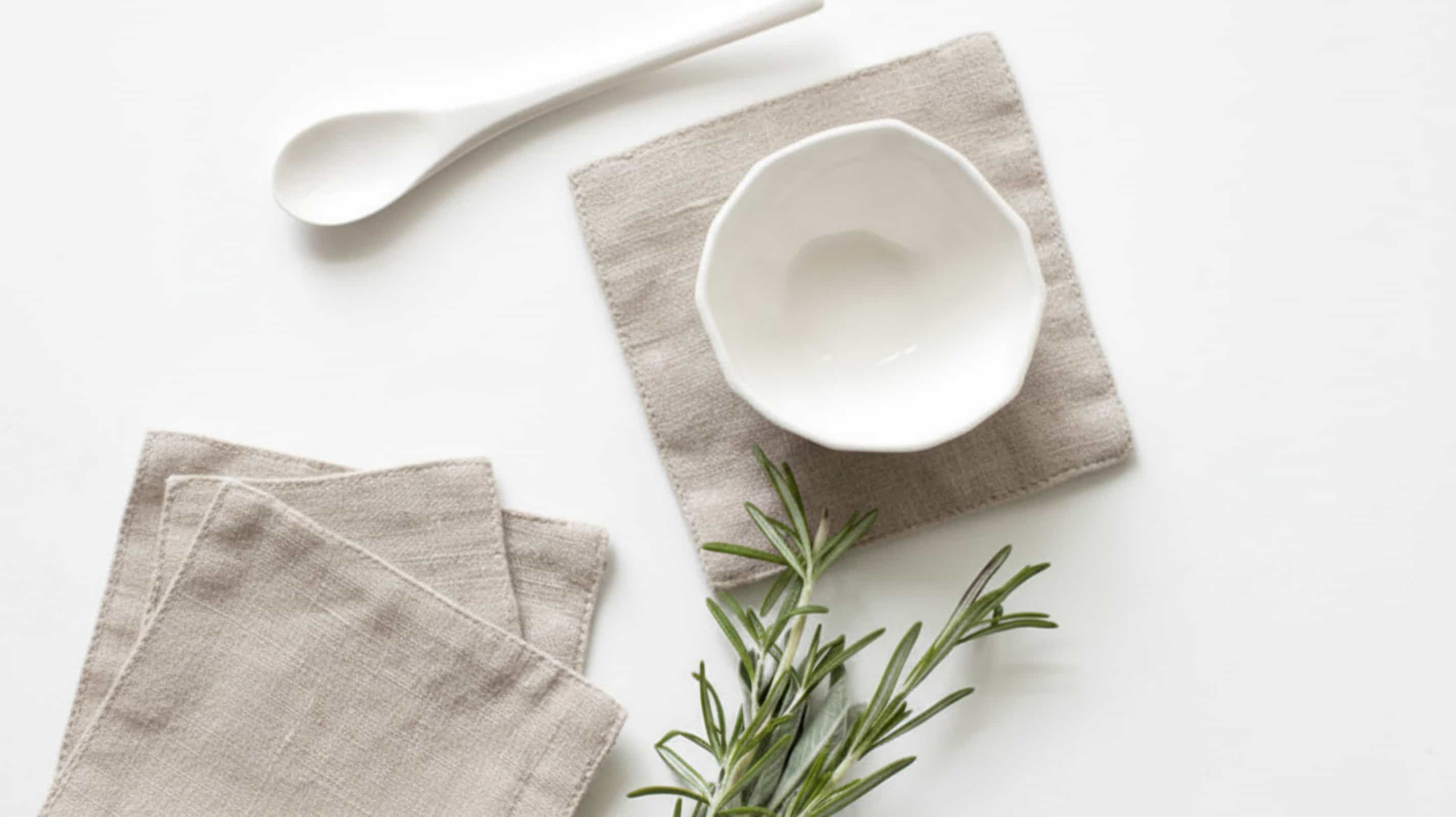 Natural linen coasters with white ceramic bowl, spoon and fresh rosemary on white background, flat lay