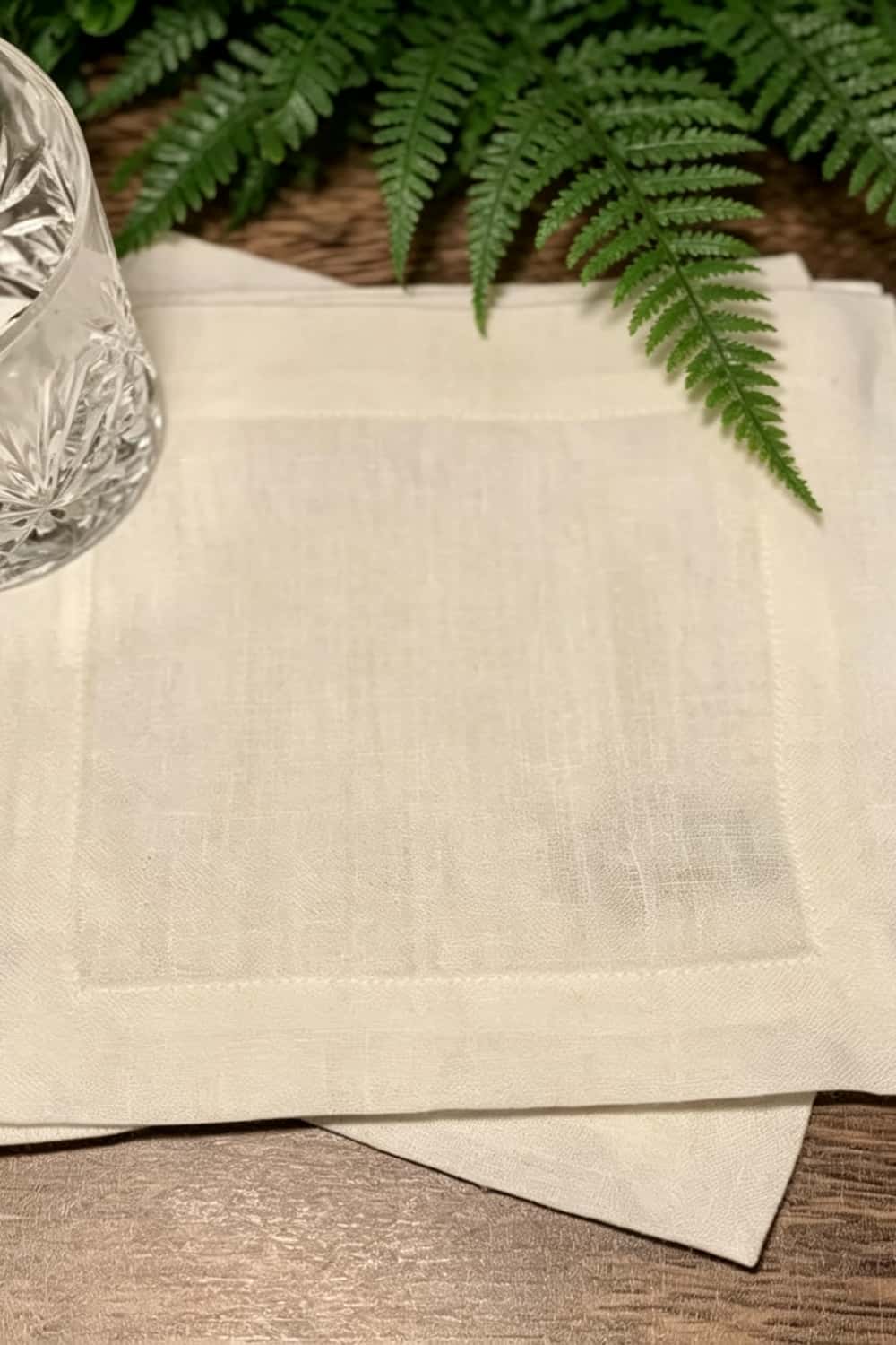 A close-up of a premium cream linen cocktail napkin featuring a crisp border stitch, styled with a crystal glass and a green fern branch, highlighting material consistency for repeat embroidery production runs.