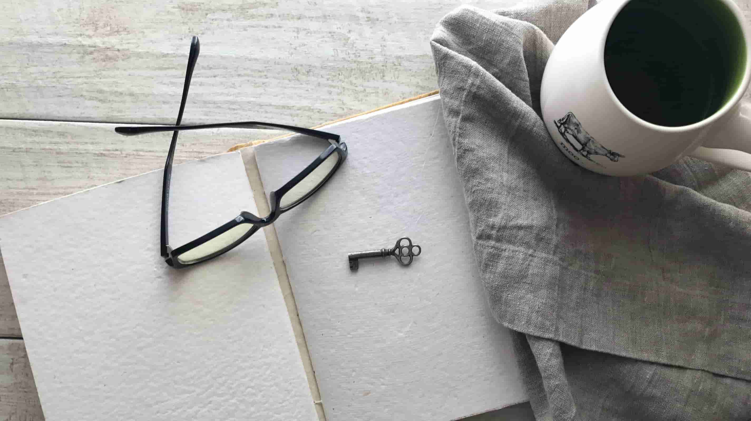 Grey washed linen napkin folded beside an open notebook, black-rimmed glasses, antique key and white coffee mug on a whitewashed wood table