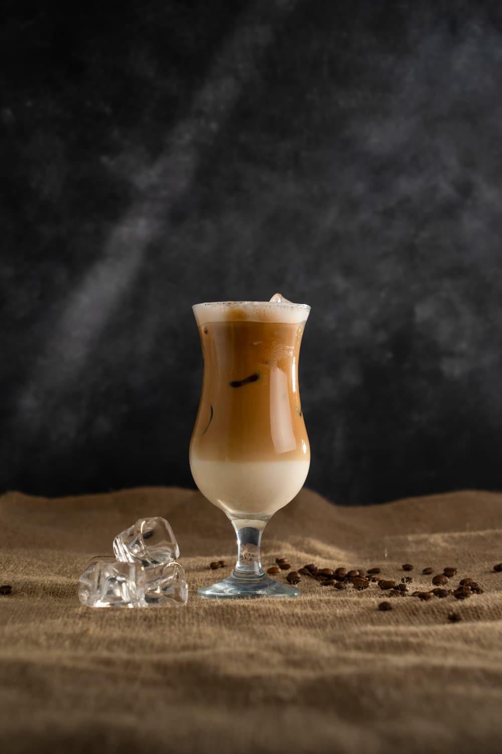 Layered iced latte in a hurricane glass with coffee beans and ice cubes on a natural burlap linen surface against a dark textured background