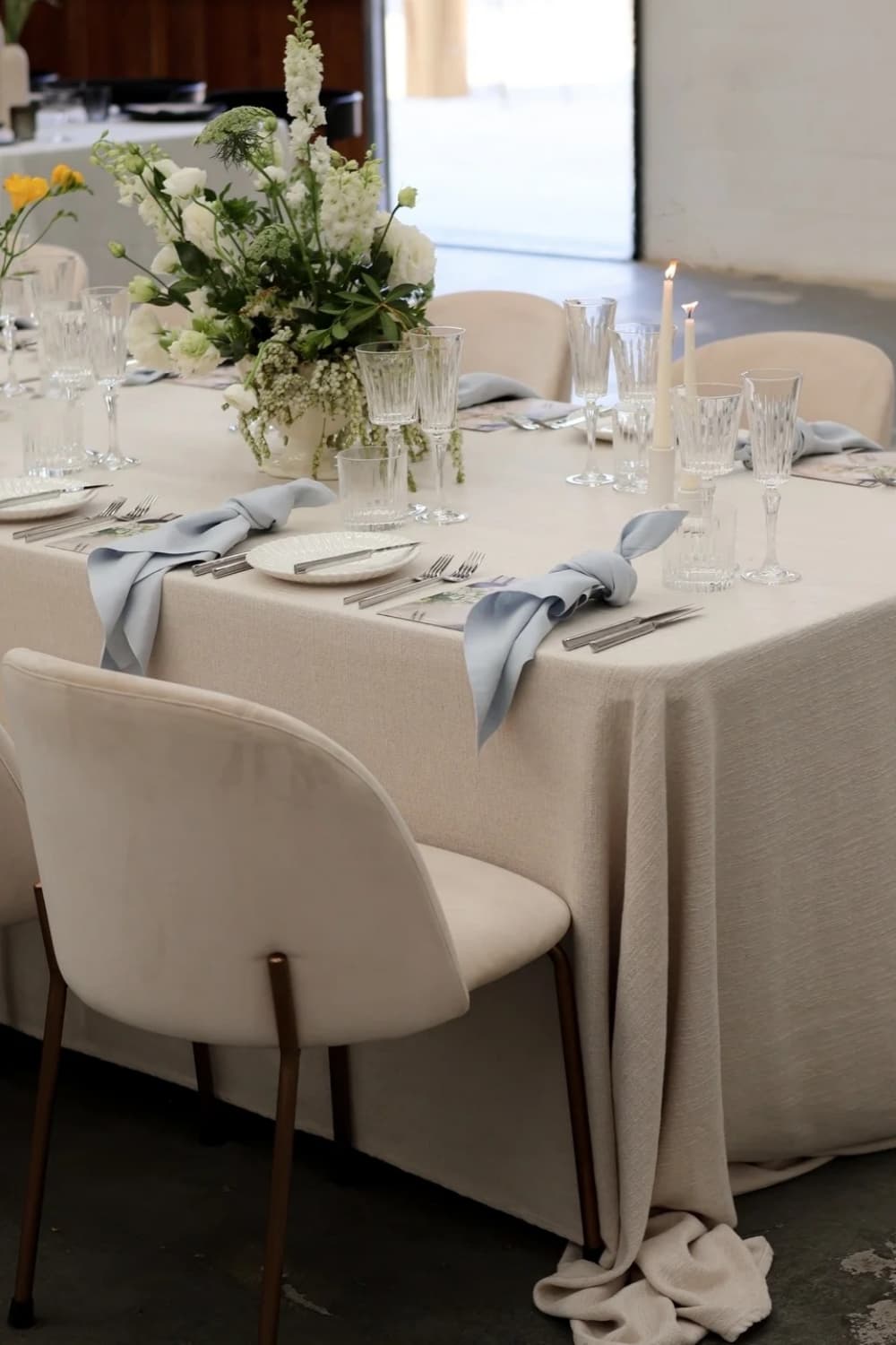 Natural beige linen tablecloth with dusty blue linen napkins, white floral centerpiece and taper candles on an oval wedding reception table