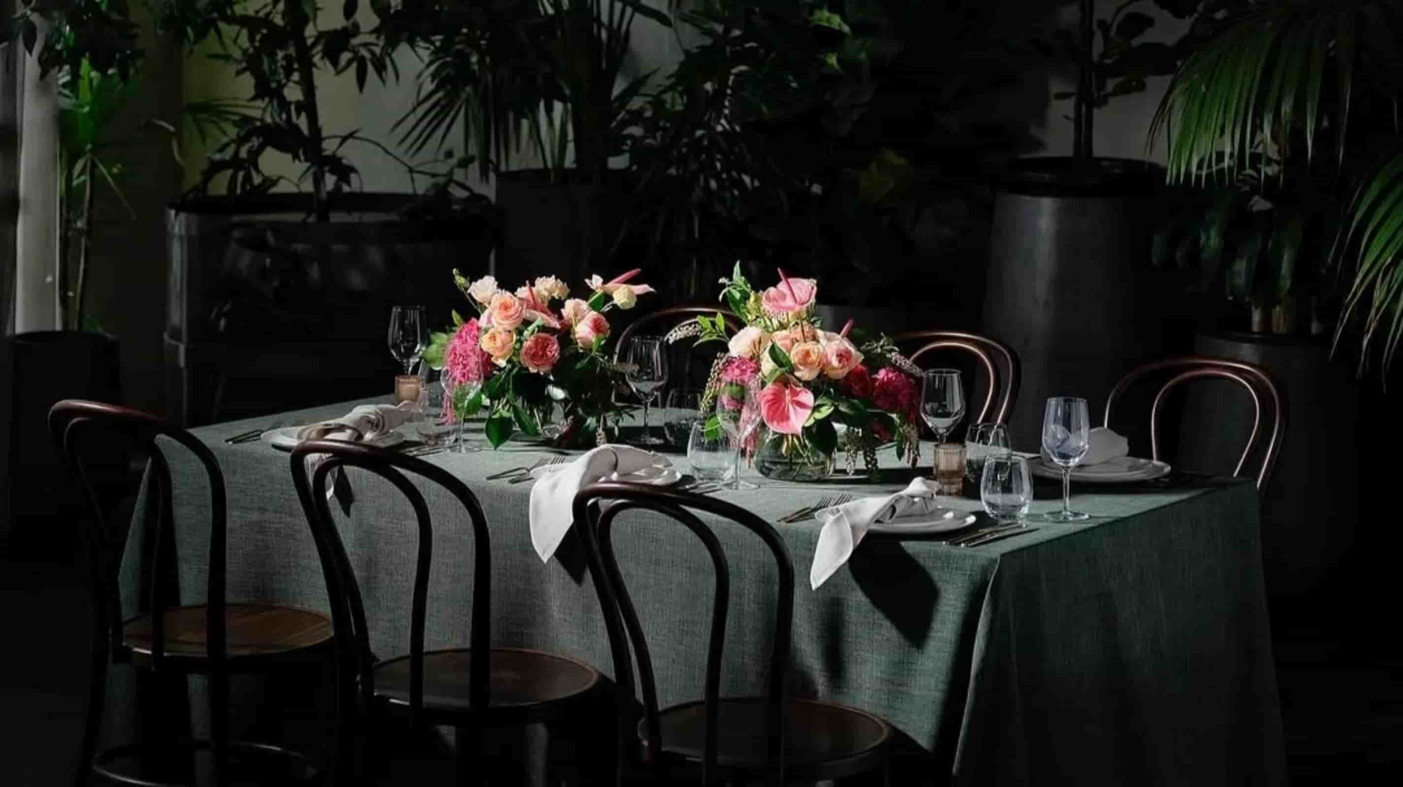Sage green washed linen tablecloth on a rectangular dining table with pink floral centerpiece and bentwood chairs in a dark botanical restaurant