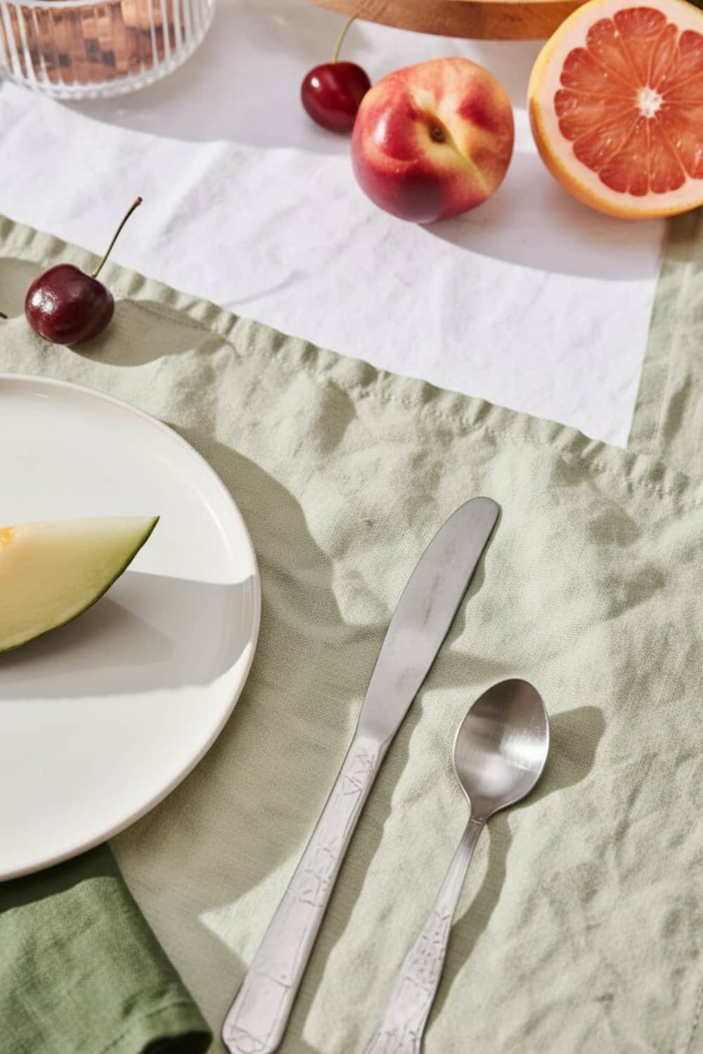 Close-up of sage green washed linen placemat with silver cutlery, white plate and fresh summer fruits