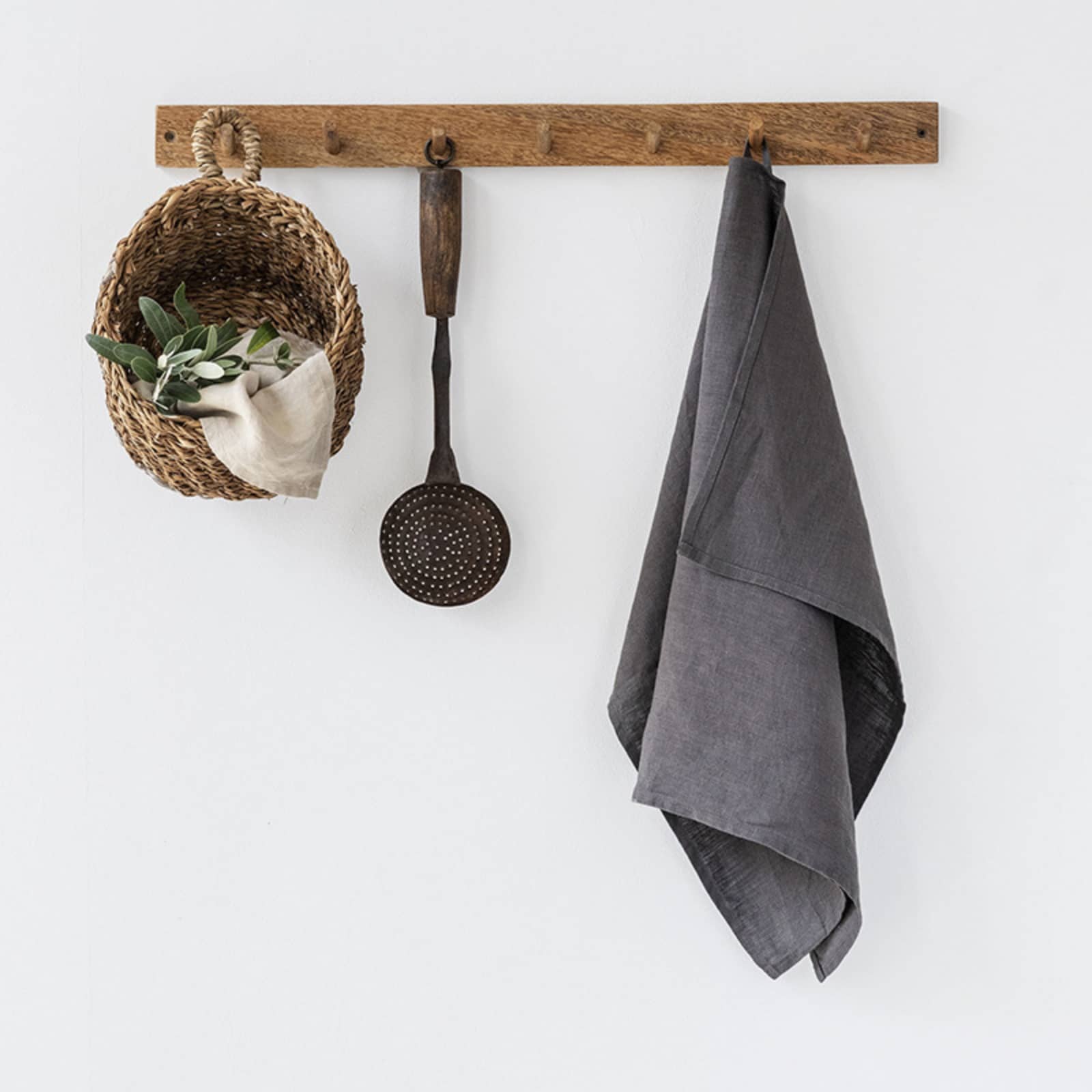 Dark grey washed linen tea towel hanging on rustic wooden hook rack with woven basket, olive branches and vintage ladle