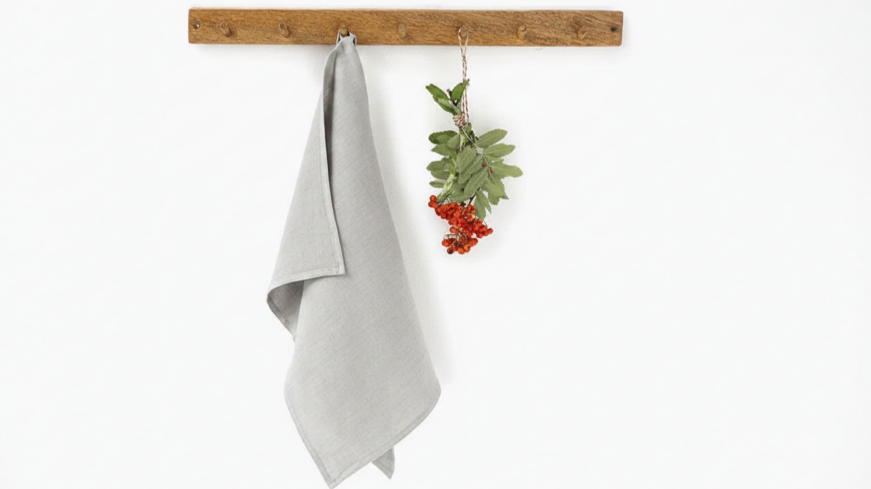 Light grey washed linen tea towel hanging on rustic wooden hook rack with red berries, white background 