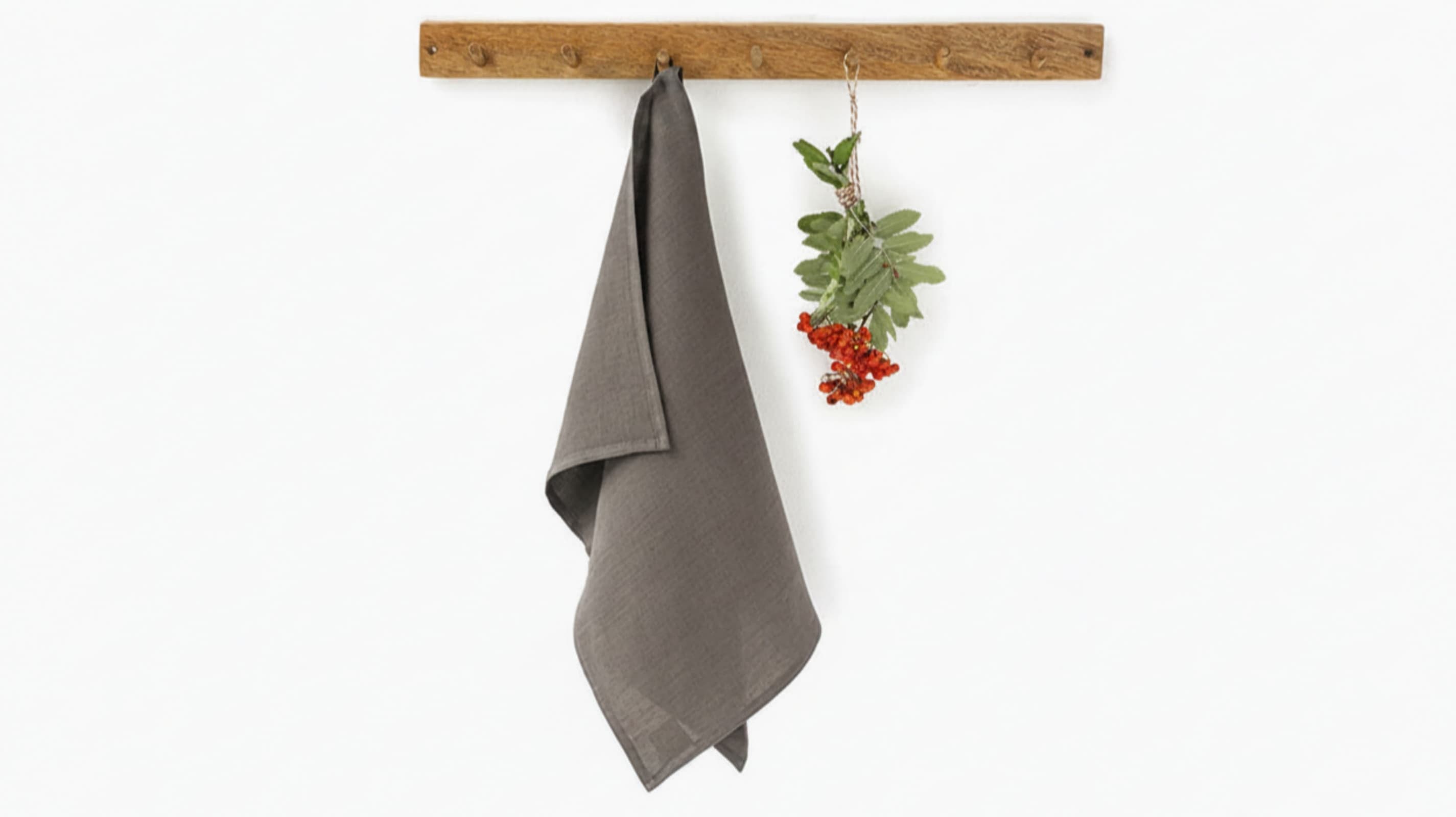 Middle grey washed linen tea towel hanging on rustic wooden hook rack with red berries, white background