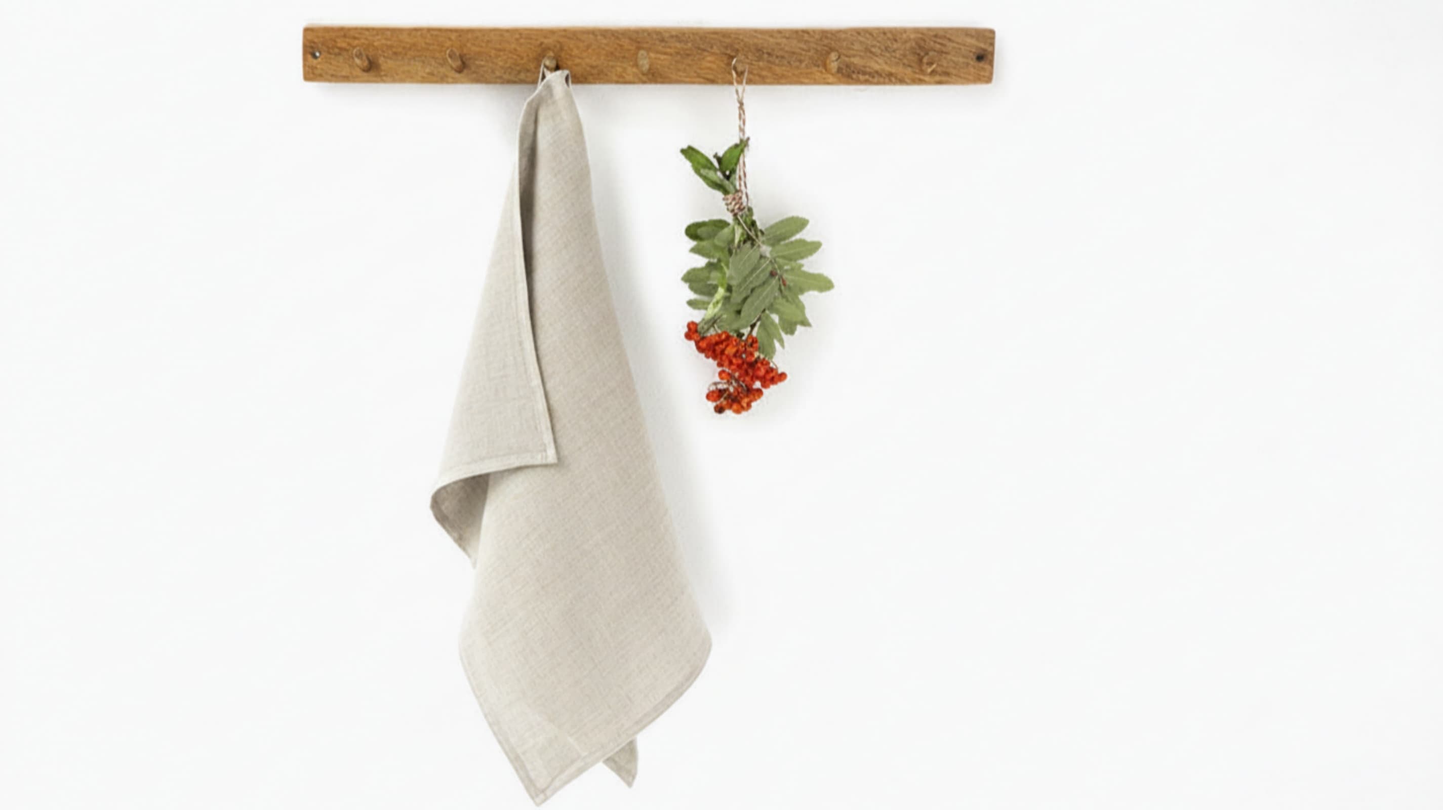 Natural linen tea towel hanging on rustic wooden hook rack with red berries, white background