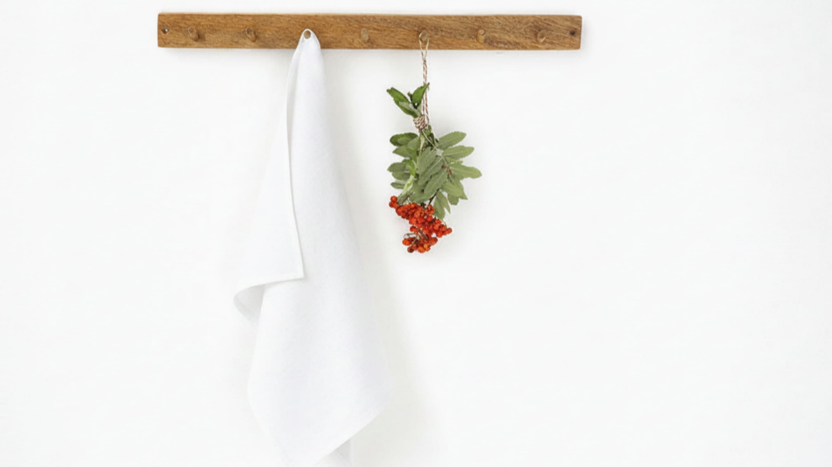 White washed linen tea towel hanging on rustic wooden hook rack with red berries, white background