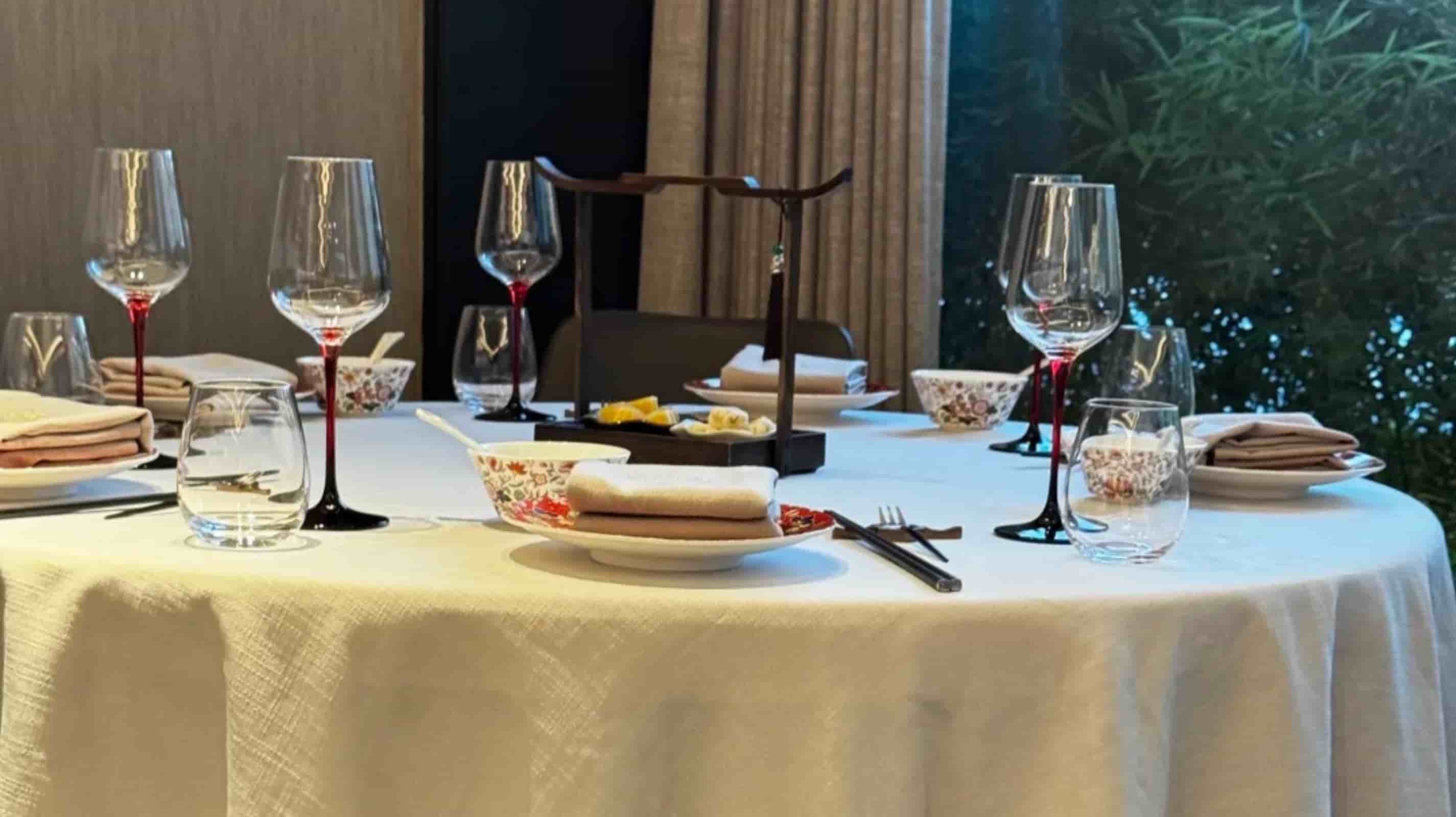 White linen round tablecloth on a large dining table set with chopsticks, floral porcelain bowls and red-stemmed wine glasses in a modern Chinese restaurant