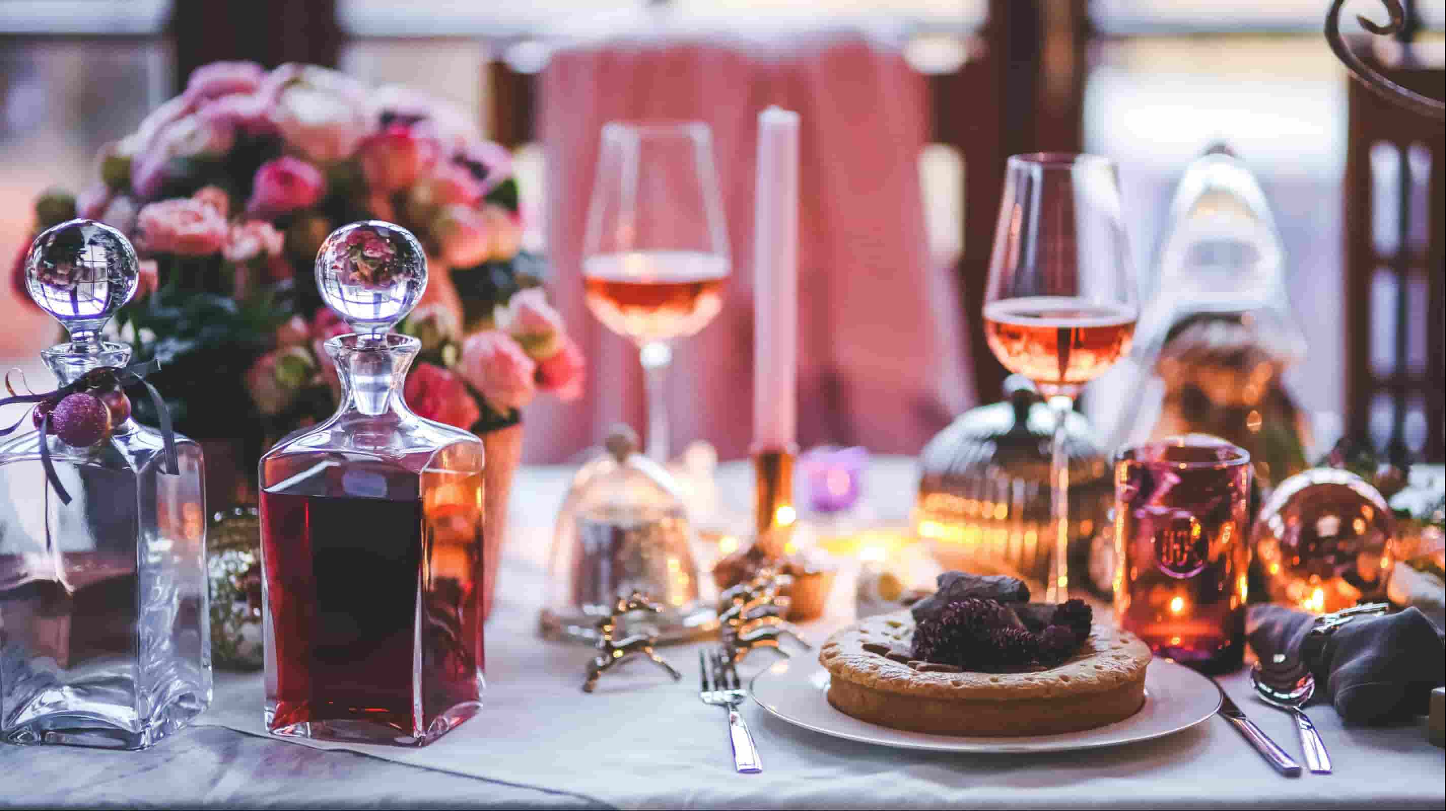 White linen tablecloth close-up with crystal decanters, rose wine glasses, taper candles and pink floral arrangement at a romantic fine dining table