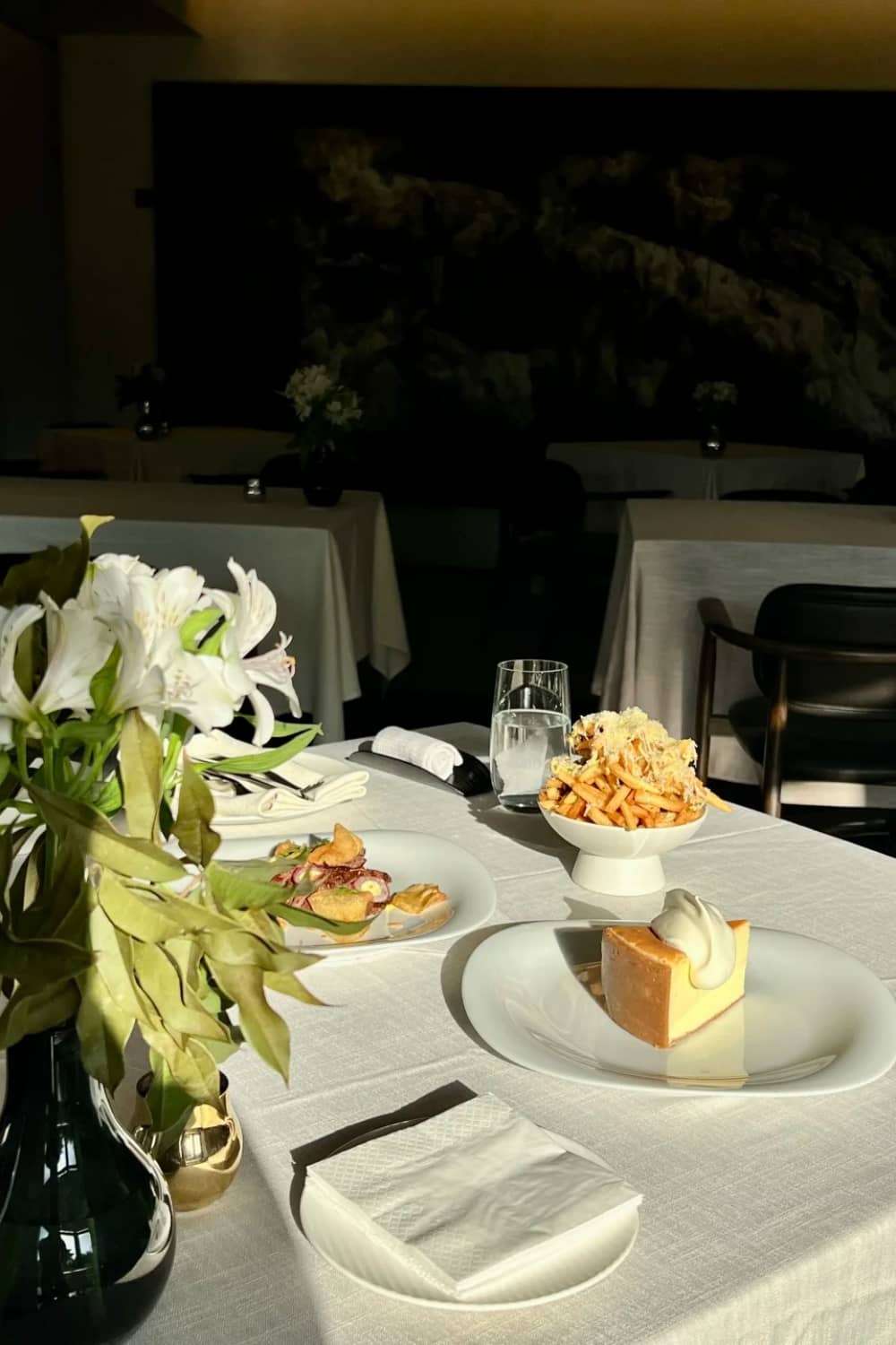 White linen tablecloth set for dinner with white lily floral arrangement, linen napkins, and gourmet dishes in a dimly lit upscale restaurant